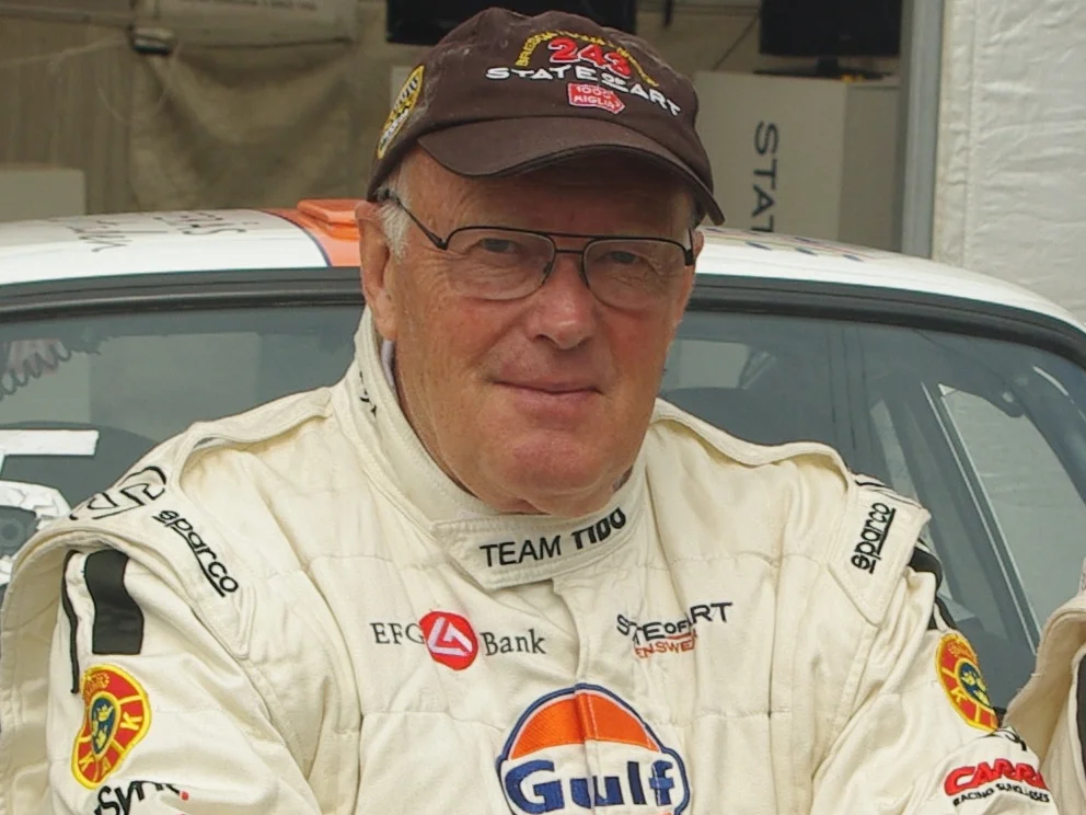 Björn Waldegård i vit tävlingsoverall och keps framför en rallybil, 2011