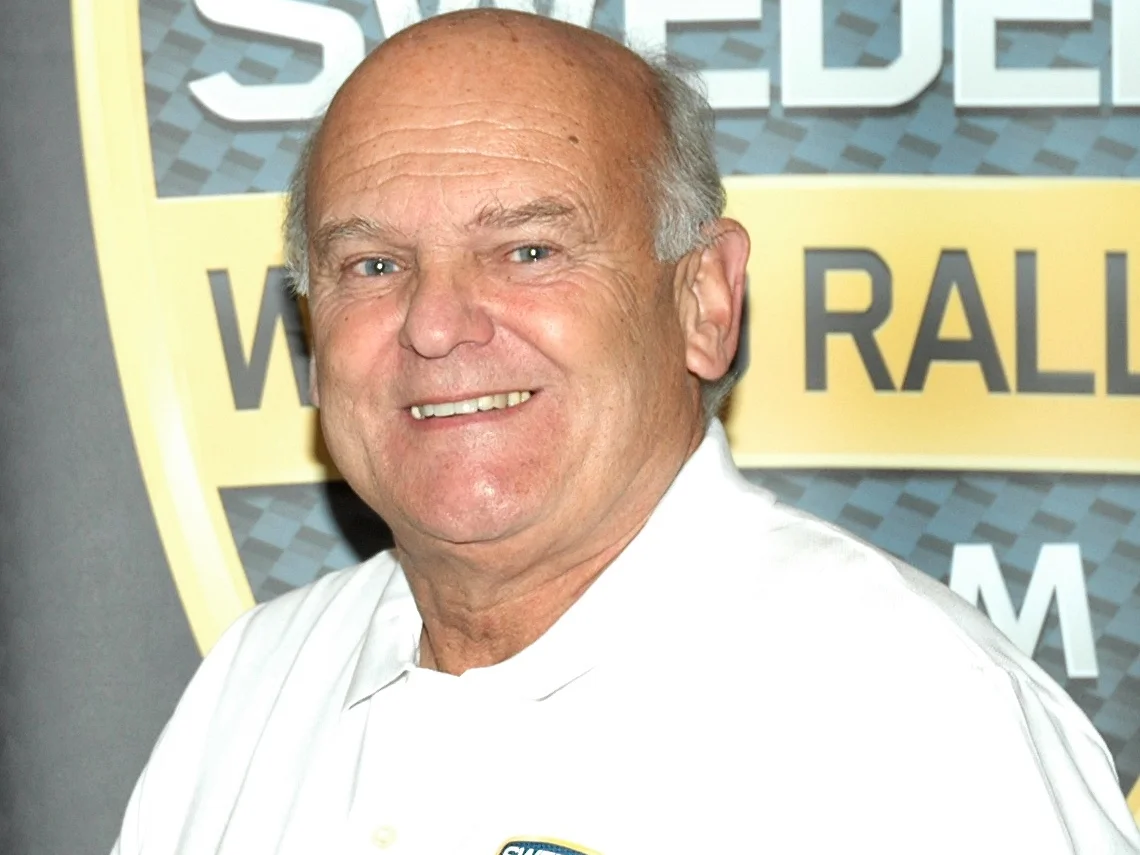 Stig Blomqvist i vit skjorta framför en rallyskylt, 2012